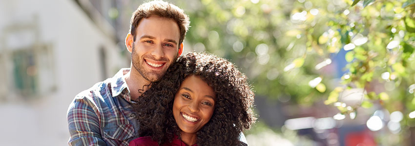 Portrait of a smiling young man hugging his girlfriend from behind while standing together in the city on a sunny day