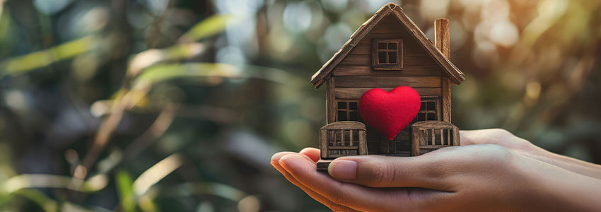 Hands holding miniature house with a red heart outdoors on a sunny summer day